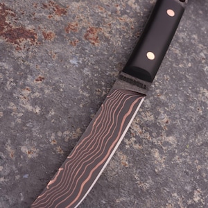 Peut inclure: Un couteau artisanal avec un manche noir et une lame pr&eacute;sentant un motif ondul&eacute; unique dans des tons de marron et de cuivre. Le manche comporte deux rivets en laiton. Une lani&egrave;re en cuir est attach&eacute;e.