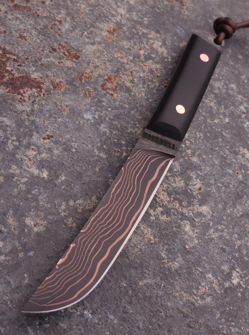 Peut inclure: Un couteau artisanal avec un manche noir et une lame pr&eacute;sentant un motif ondul&eacute; unique dans des tons de marron et de cuivre. Le manche comporte deux rivets en laiton. Une lani&egrave;re en cuir est attach&eacute;e.