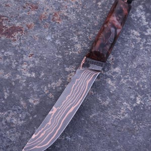 Peut inclure: Un couteau artisanal avec une lame &agrave; motifs et un manche avec un design marbr&eacute; marron et blanc. La lame pr&eacute;sente un motif unique de grain de bois. Un cordon noir est attach&eacute; &agrave; la poign&eacute;e.