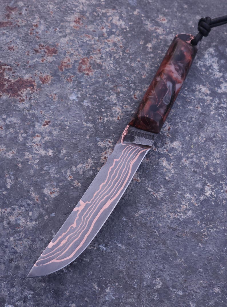 Peut inclure: Un couteau artisanal avec une lame &agrave; motifs et un manche avec un design marbr&eacute; marron et blanc. La lame pr&eacute;sente un motif unique de grain de bois. Un cordon noir est attach&eacute; &agrave; la poign&eacute;e.