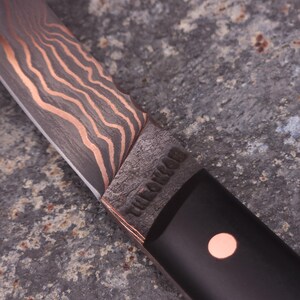 Peut inclure: Gros plan sur un couteau artisanal avec une lame &agrave; motifs et des accents cuivr&eacute;s. Le manche est noir avec un point cuivr&eacute;. La lame porte l'inscription "THE LONEMAN".