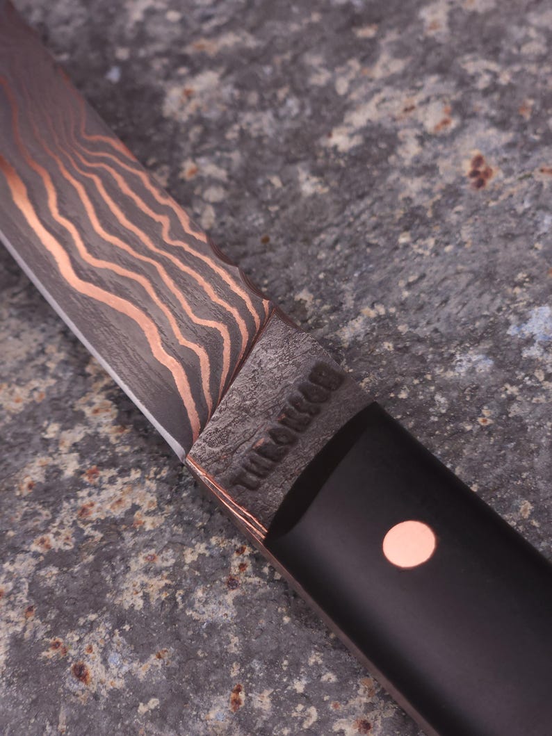 Peut inclure: Gros plan sur un couteau artisanal avec une lame &agrave; motifs et des accents cuivr&eacute;s. Le manche est noir avec un point cuivr&eacute;. La lame porte l'inscription "THE LONEMAN".