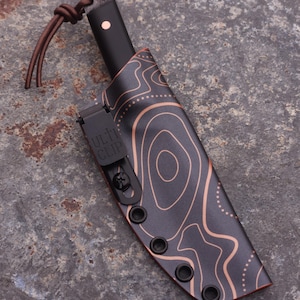 Peut inclure: Un couteau noir avec un &eacute;tui &agrave; motif topographique cuivr&eacute;. Le manche du couteau est noir avec un point cuivr&eacute;. L'&eacute;tui a un clip noir avec le texte "ULTI CLIP". Une lani&egrave;re en cuir marron est attach&eacute;e au couteau.