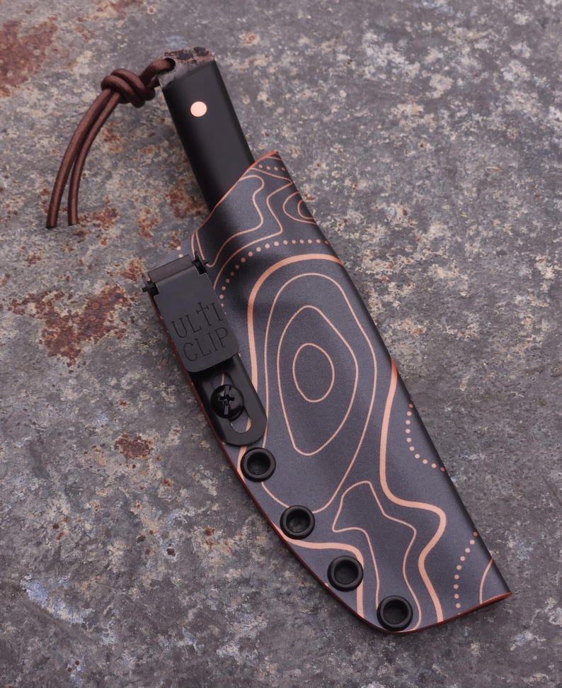 Peut inclure: Un couteau noir avec un &eacute;tui &agrave; motif topographique cuivr&eacute;. Le manche du couteau est noir avec un point cuivr&eacute;. L'&eacute;tui a un clip noir avec le texte "ULTI CLIP". Une lani&egrave;re en cuir marron est attach&eacute;e au couteau.