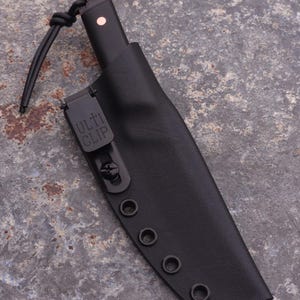Peut inclure: Un couteau noir avec un &eacute;tui en cuir noir. L'&eacute;tui comporte un clip m&eacute;tallique avec le texte "ULTI CLIP" et plusieurs &oelig;illets. Le manche du couteau a un petit accent rond rose. Un cordon noir est attach&eacute;.