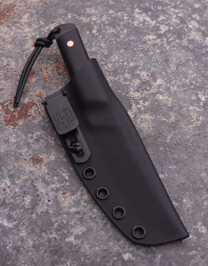Peut inclure: Un couteau noir avec un &eacute;tui en cuir noir. L'&eacute;tui comporte un clip m&eacute;tallique avec le texte "ULTI CLIP" et plusieurs &oelig;illets. Le manche du couteau a un petit accent rond rose. Un cordon noir est attach&eacute;.