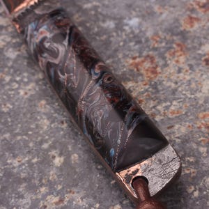 Peut inclure: Gros plan sur un manche de couteau avec un motif marbr&eacute; dans les tons de marron, noir et bleu. La poign&eacute;e est orn&eacute;e de m&eacute;tal couleur cuivre et d'une lani&egrave;re en cuir marron.