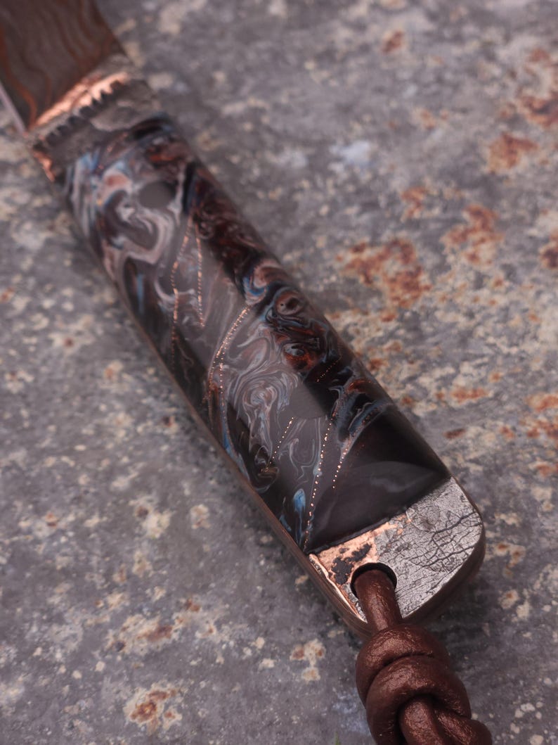 Peut inclure: Gros plan sur un manche de couteau avec un motif marbr&eacute; dans les tons de marron, noir et bleu. La poign&eacute;e est orn&eacute;e de m&eacute;tal couleur cuivre et d'une lani&egrave;re en cuir marron.