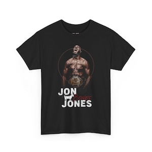 Puede incluir: Camiseta negra con un gráfico de un hombre musculoso con un cinturón de la UFC, dentro de un círculo marrón. El texto "JON JONES" se muestra en blanco, con un gráfico de cabra entre los nombres. Cuello redondo.