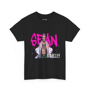 May include: Black t-shirt featuring a graphic of a person with tattoos, wearing a white robe, and green shorts. The name "SEAN" is in pink graffiti-style lettering above the person, with "O'MALLEY" below.
