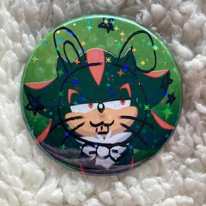 Puede incluir: Un botón verde y negro con una imagen de dibujos animados de Shadow the Hedgehog vestido de gato. El botón tiene un acabado brillante y estrellas en el fondo.