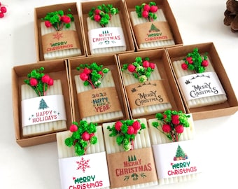 May include: A collection of nine rectangular white soaps, each individually packaged in a brown cardboard box. Each soap is wrapped with a paper band featuring various holiday-themed graphics and text such as Merry Christmas and Happy Holidays, topped with small sprigs of green and red dried flowers.