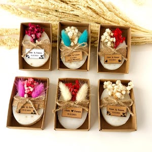 May include: Six oval-shaped soaps, each in a small cardboard box, are decorated with dried flowers and twine. Each soap has a small tag with the names "Liam & Emma" and the date "12.12.2024". The flowers are in various colors.