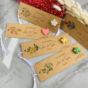 May include: Kraft paper bookmarks with floral designs and the text "LET LOVE GROW" along with names and a date. Each bookmark has a white tassel and a small decorative element, such as a flower or heart.