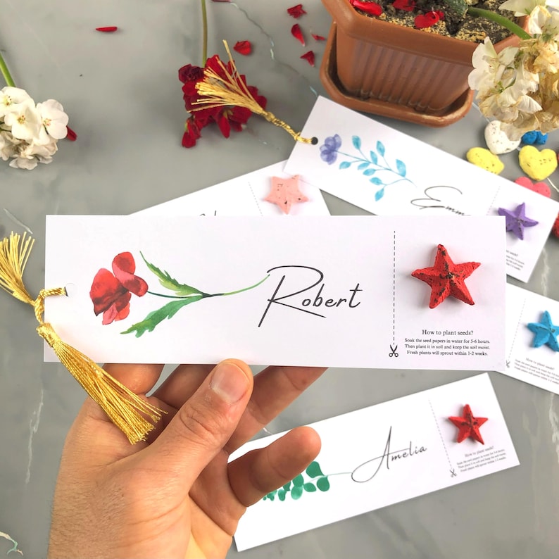 May include: White rectangular seed paper bookmarks with floral designs and names. Each bookmark features a colourful star-shaped seed pod. One bookmark has the name "Robert" and instructions on how to plant seeds.