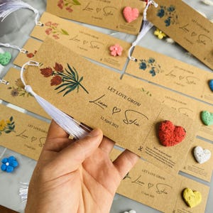 May include: Kraft paper bookmarks with floral designs and the text "LET LOVE GROW" and names "Liam & Sofia" with a date. Each bookmark has a white tassel and a small heart-shaped seed packet.