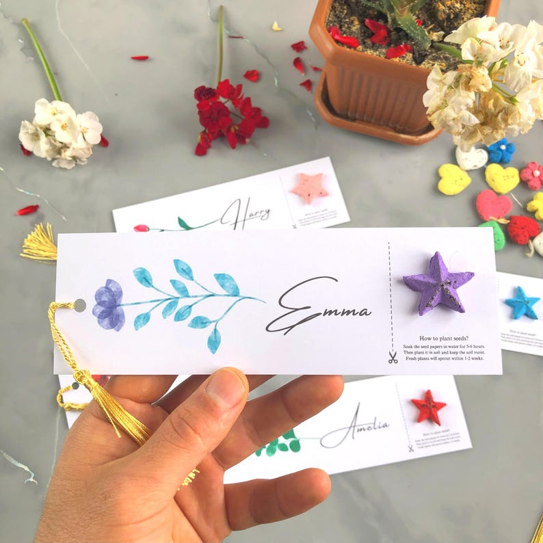 May include: A white bookmark featuring a watercolour floral design and the name "Emma" in cursive. A purple star-shaped seed paper is attached. The bookmark has a gold tassel and planting instructions.
