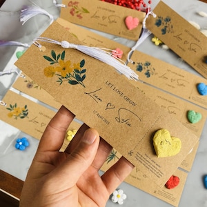 May include: Kraft paper bookmark with a white tassel, yellow floral design, and the text "LET LOVE GROW Liam & Sofia 12 April 2025." Includes a heart-shaped seed packet. Other bookmarks and decorative hearts are in the background.