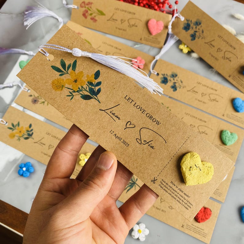 May include: Kraft paper bookmark with a white tassel, yellow floral design, and the text "LET LOVE GROW Liam & Sofia 12 April 2025." Includes a heart-shaped seed packet. Other bookmarks and decorative hearts are in the background.
