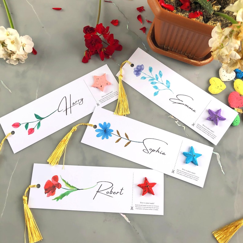 May include: Four white bookmarks with floral designs and gold tassels. Each bookmark features a name: Harry, Emma, Sophia, and Robert. Each bookmark has a small star attached. The bookmarks are on a grey surface.