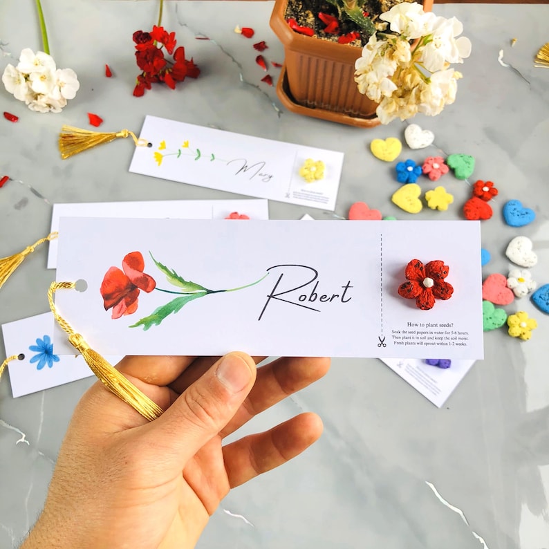 May include: A hand holding a white bookmark with the name "Robert" in script. The bookmark features a red watercolor flower and a gold tassel. Other bookmarks and colourful flower-shaped decorations are visible.