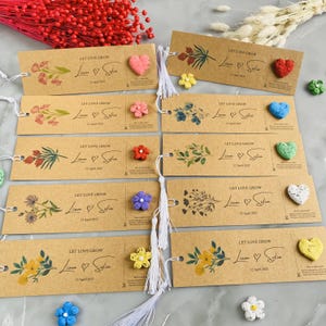 May include: A collection of brown paper bookmarks with floral designs and the text "LET LOVE GROW Liam & Sofia 12 April 2025". Each bookmark has a small, colorful, heart-shaped or flower-shaped embellishment. White tassels are attached.