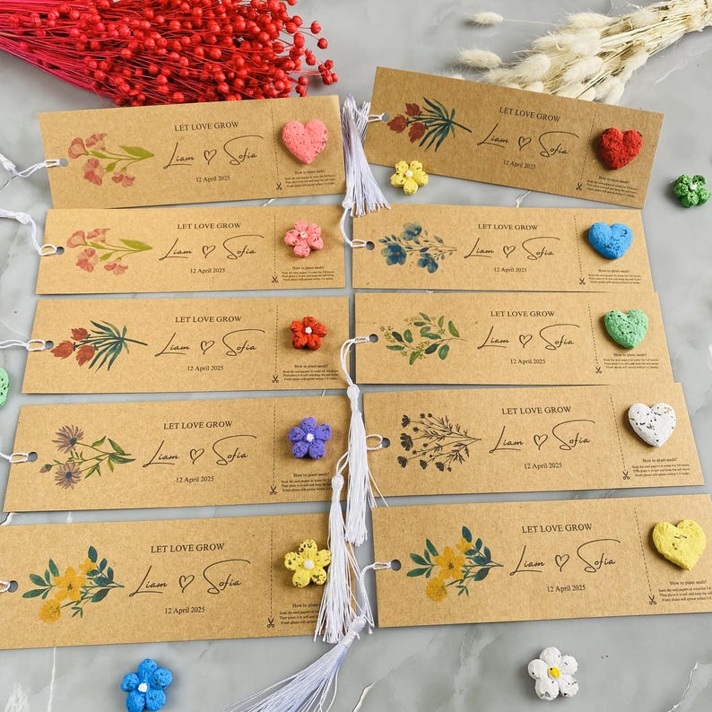 May include: A collection of brown paper bookmarks with floral designs and the text "LET LOVE GROW Liam & Sofia 12 April 2025". Each bookmark has a small, colorful, heart-shaped or flower-shaped embellishment. White tassels are attached.