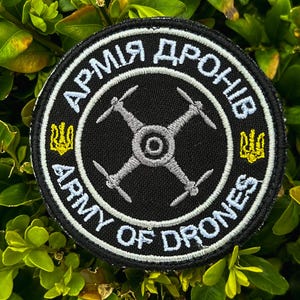 May include: A black and white embroidered patch with the text "Army of Drones" in white and "Армія дронів" in black. The patch features a grey drone with four propellers in the centre. The patch is surrounded by a white border and a black outer border.