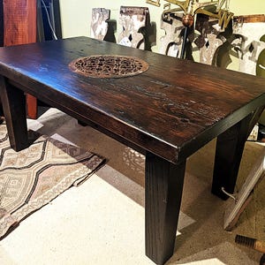 May include: A dark brown wooden table with a decorative metal insert. The table has a rectangular top and four thick, square legs. The wood grain is visible, and the table appears to be handcrafted.
