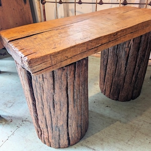May include: A rustic wooden bench with a rectangular top supported by two cylindrical log legs. The bench is made of weathered, natural wood with visible grain and knots, showcasing a warm, brown color. The design suggests a handcrafted, outdoor or farmhouse style.