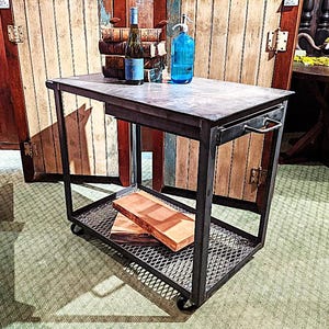 May include: A black metal bar cart with a gray countertop and a lower shelf with a diamond pattern. The cart has a handle and wheels. A bottle of wine and a blue bottle are on the top shelf. Two wooden cutting boards are on the lower shelf.