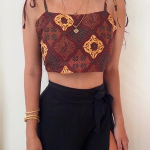 Pode incluir: Um top curto estampado em marrom e amarelo com alças finas que amarram nos ombros. O top é feito de um tecido leve e tem um decote quadrado. A modelo está usando o top com uma saia preta.