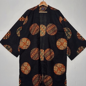 Può includere: Una veste in stile kimono nera con grandi motivi circolari nei toni del marrone, arancione e verde. La veste è appesa a una gruccia di legno su uno sfondo bianco. I cerchi presentano intricati disegni geometrici.