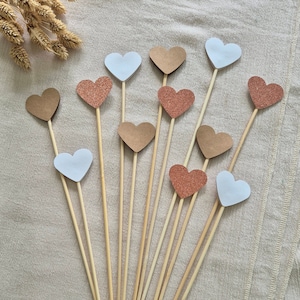 Peut inclure: Un ensemble de 12 brochettes en bois avec des sommets en forme de cœur. Les sommets sont de trois couleurs : blanc, marron et rose pailleté.