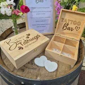 Peut inclure: Deux boîtes en bois clair avec couvercles à charnières, l'une fermée et l'autre ouverte, gravées au laser avec le texte "Le Petit Bar à Tatouages éphémères" et "TATTOO Bar". Inclut un panneau d'instructions encadré, deux éponges en forme de cœur.