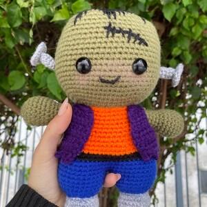 May include: A green crocheted Frankenstein monster doll with black eyes, a stitched scar, and a purple and orange waistcoat. The doll is wearing blue trousers and grey shoes.