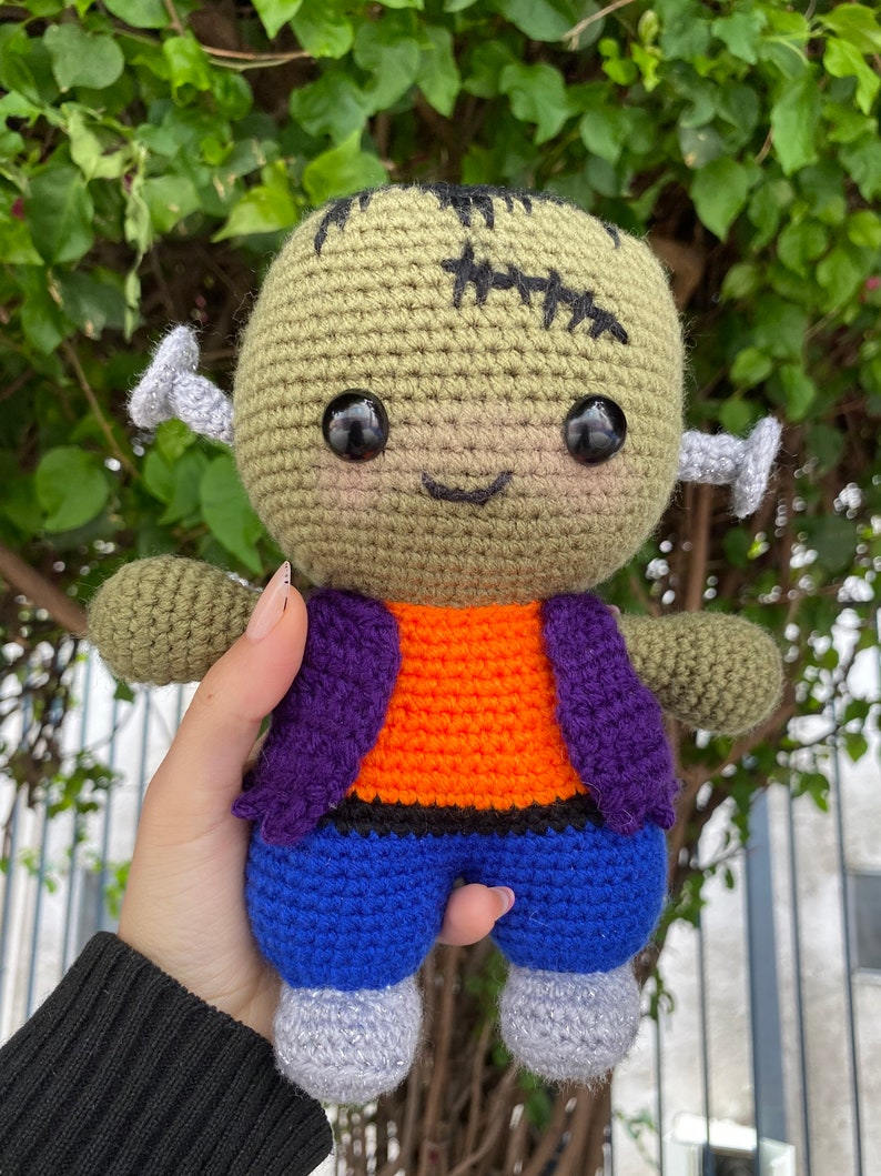 May include: A green crocheted Frankenstein monster doll with black eyes, a stitched scar, and a purple and orange waistcoat. The doll is wearing blue trousers and grey shoes.
