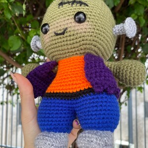 May include: A green crocheted Frankenstein monster doll with black eyes, a purple and orange shirt, blue trousers, and silver glitter shoes.