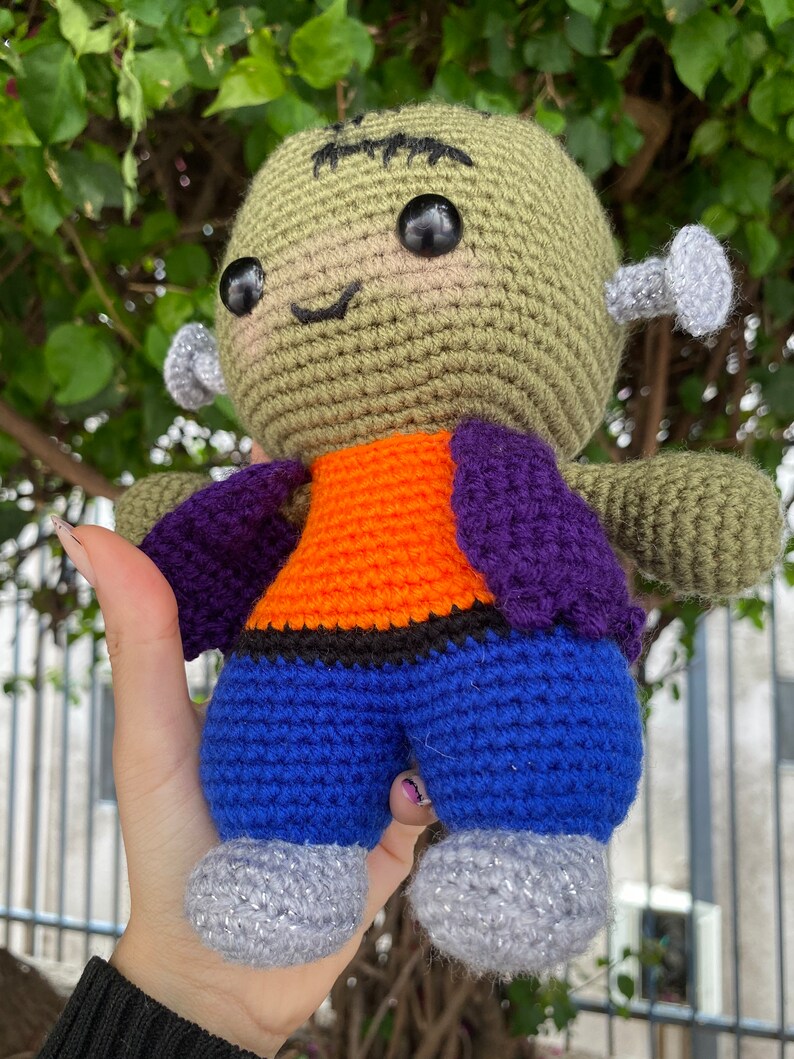 May include: A green crocheted Frankenstein monster doll with black eyes, a purple and orange shirt, blue trousers, and silver glitter shoes.