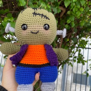 May include: A green crocheted Frankenstein monster doll with black stitches, orange and purple clothing, and blue trousers. The doll has large black eyes and a stitched-on smile.