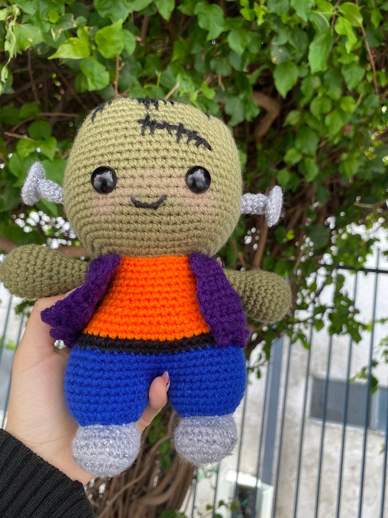May include: A green crocheted Frankenstein monster doll with black stitches, orange and purple clothing, and blue trousers. The doll has large black eyes and a stitched-on smile.