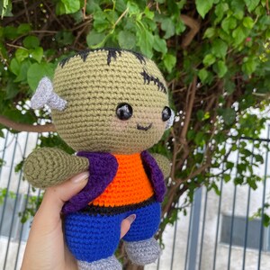 May include: A green crocheted Frankenstein monster plush toy with purple and orange clothing and blue trousers. The toy has black eyes and a stitched scar on its forehead.