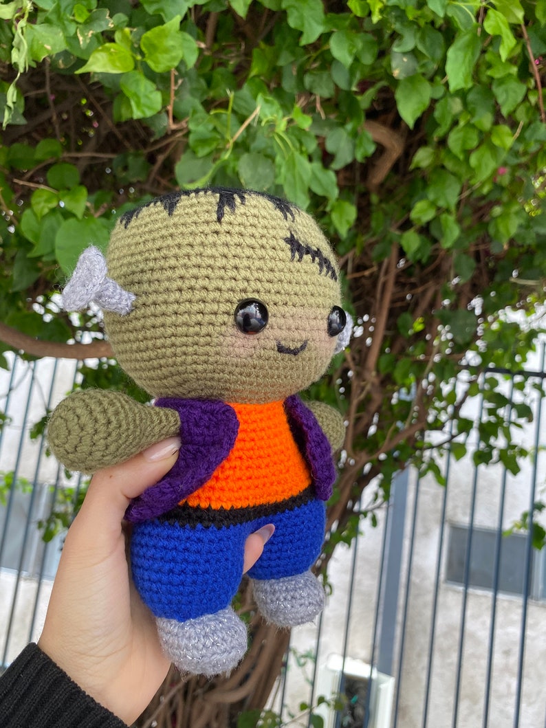 May include: A green crocheted Frankenstein monster plush toy with purple and orange clothing and blue trousers. The toy has black eyes and a stitched scar on its forehead.