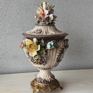 Könnte beinhalten: Dekorative cremefarbene Keramikurne mit Deckel und bunten Blumenverzierungen. Die Urne hat einen Sockel und einen goldfarbenen Fuß. Der Deckel ist mit Blumen gekrönt. Ein dekorativer Artikel für das Zuhause.