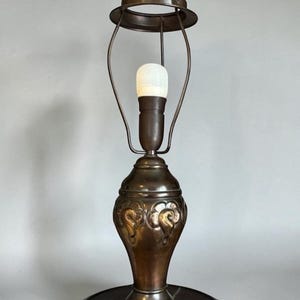 May include: A bronze table lamp with a decorative floral design on the base. The lamp has a bulb and a circular shade holder. It is set on a dark wooden round base.