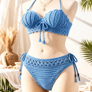 May include: A light blue crochet bikini set displayed on a mannequin. The top features a seashell-like design with halter straps and tie closures. The bottoms have a matching crochet pattern with tie sides. A gold necklace is visible.