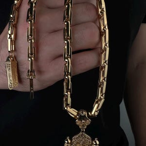 May include: A gold-colored necklace with a large pendant featuring an ornate religious design. The chain is made of thick, rectangular links. The pendant depicts a religious figure and architectural details.