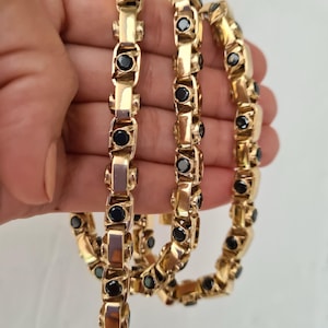 May include: A gold chain necklace with black gemstones. The chain is made of interlocking links with a square shape. The gemstones are set into the links and are round in shape.