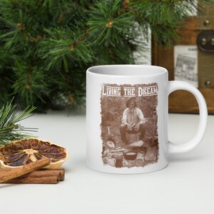 Puede incluir: Taza de cerámica blanca con una imagen marrón de un hombre sentado en un entorno de bosque. El texto "LIVING THE DREAM" está impreso en marrón en la taza.
