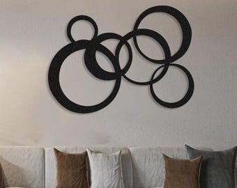 May include: A modern living room featuring a white sectional sofa with several throw pillows in shades of brown and gray. A large black metal wall art piece with overlapping circles hangs above the sofa. A green plant and a black lamp are also visible.